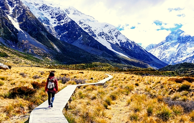 Woman walking on boardwalk across field near mountains